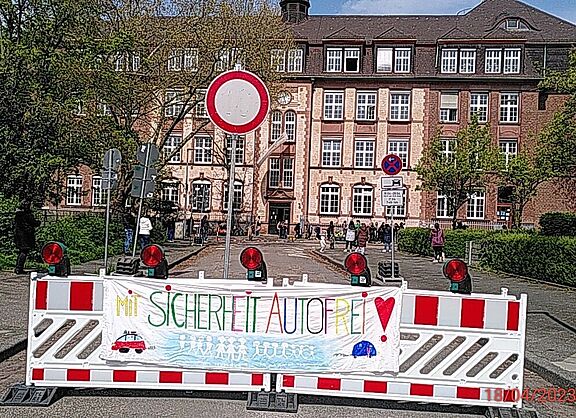 Das Bild zeigt die Sperre an der Schulstraße mit einem von Kindern gemalten Transparent mit der Aufschrift 'Mit Sicherheit Autofrei'
