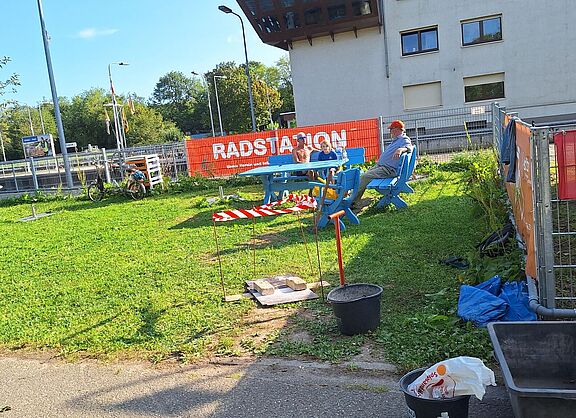 Das Bild zeigt den Radknoten mit drei Besuchern, die auf einer Bank sitzen, im Vordergrund der Ort an dem die Radservicestation plaziert werden soll.