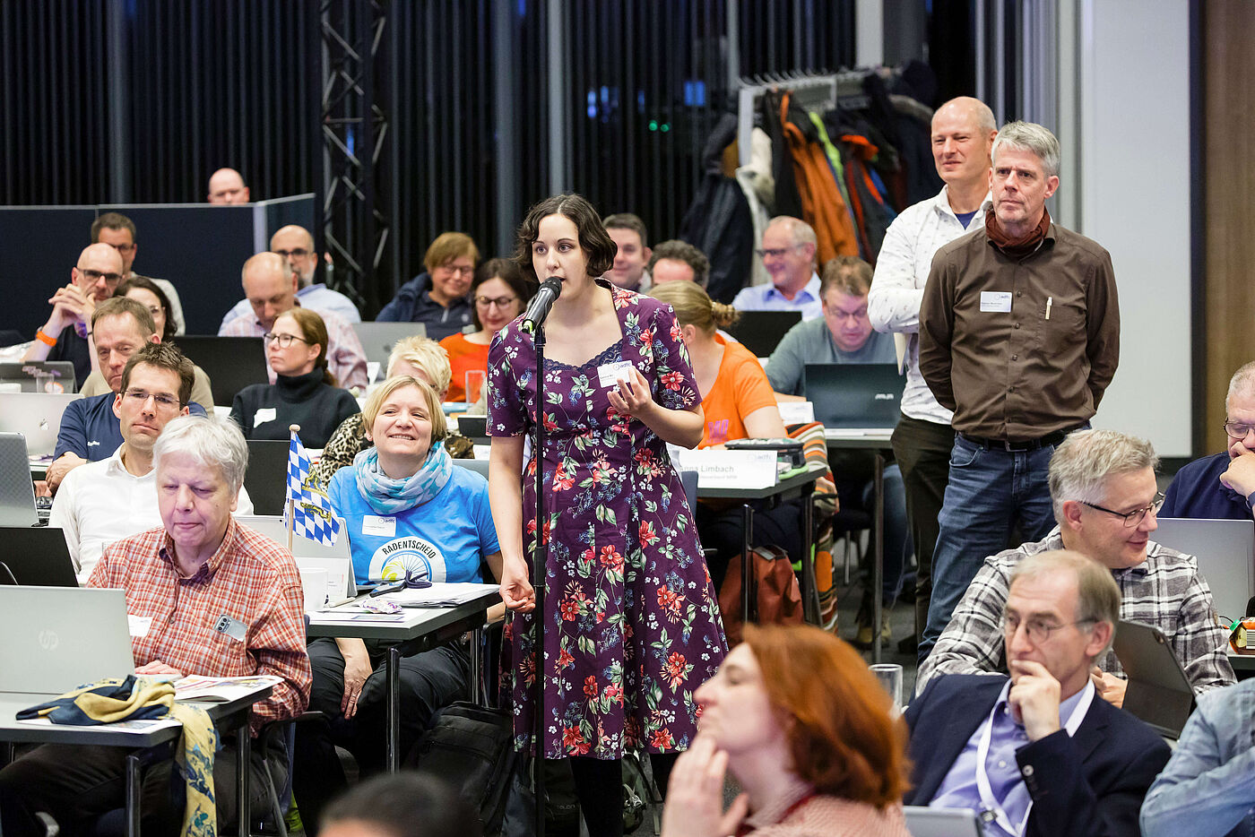 Eine Frau am Mikrofon in einem Saal voller Menschen.